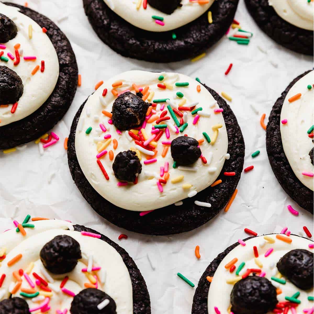 Homemade Oreo cookies topped with white cake batter frosting, sprinkles, and Oreo truffles on a white background.