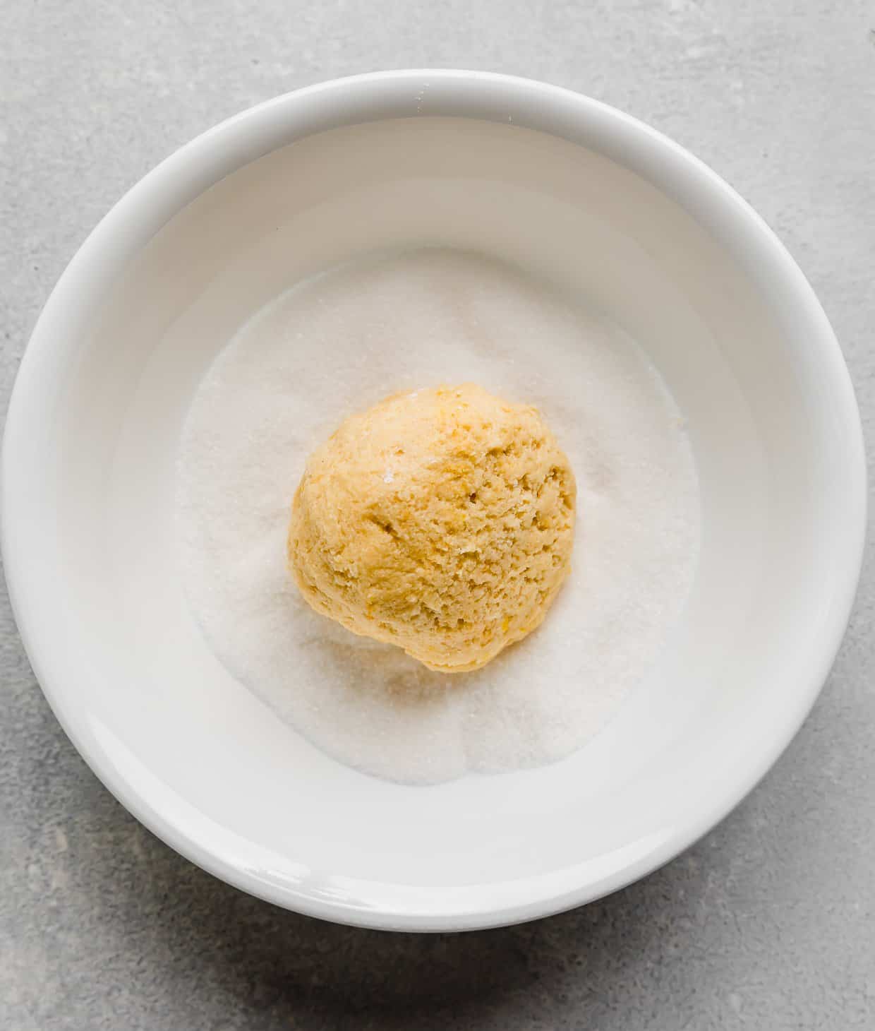 Lemon Crinkles cookie dough in a white bowl filled with granulated sugar.
