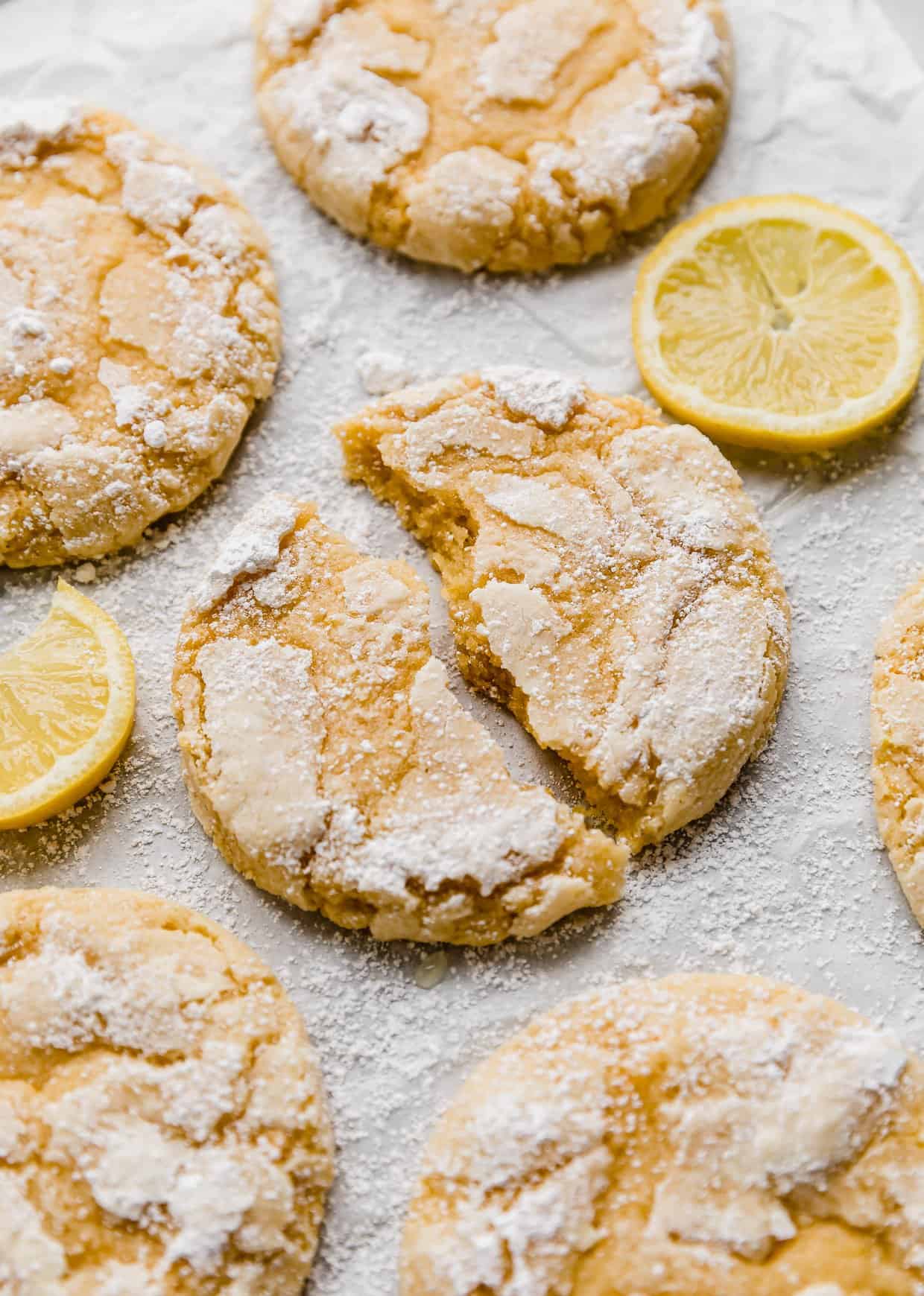 A Lemon Crinkle Cookie torn in half, on a white background with lemon slices around the cookie.