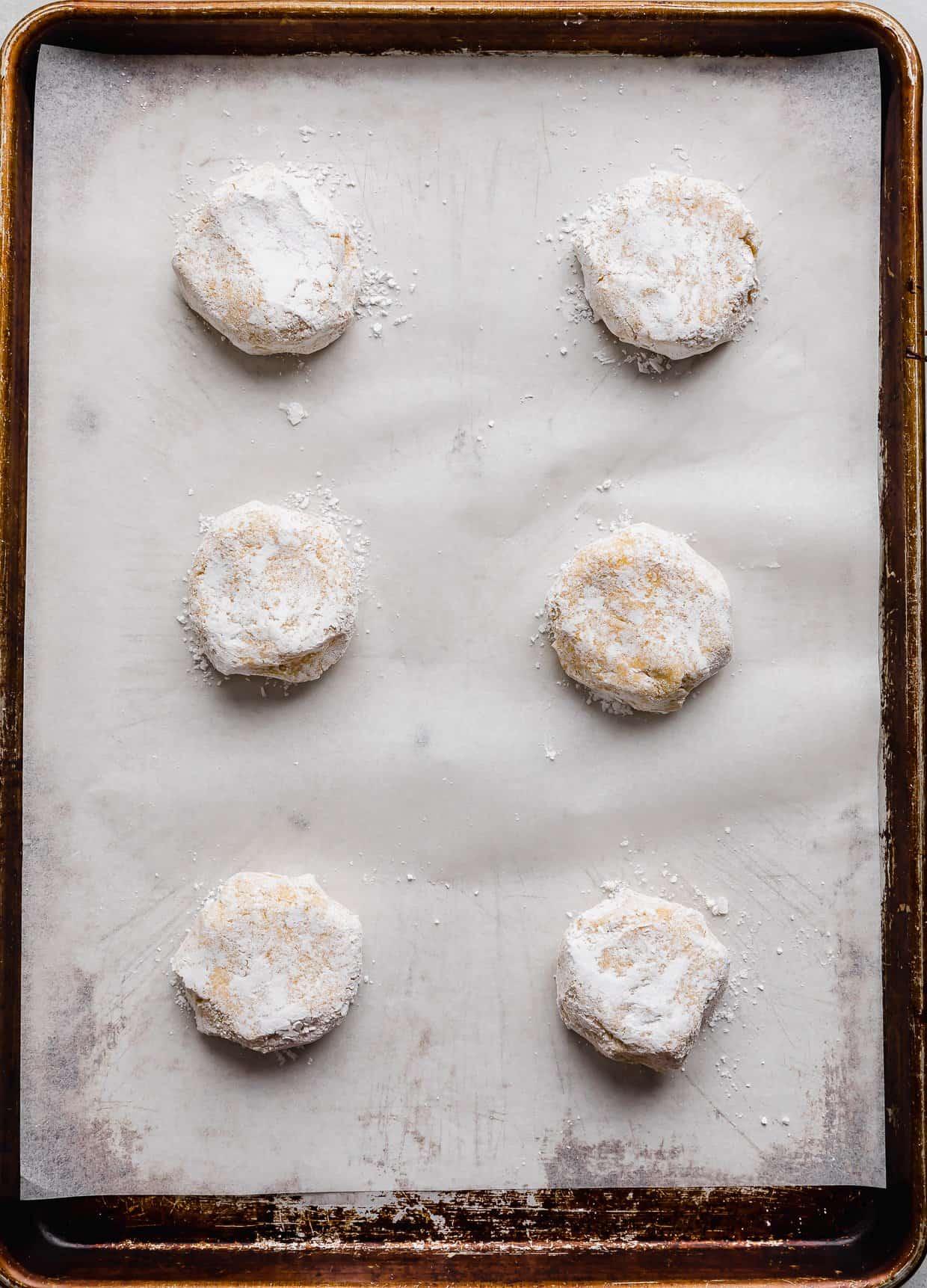Six lemon crinkle cookie dough balls rolled in powdered sugar on a white parchment lined baking sheet.