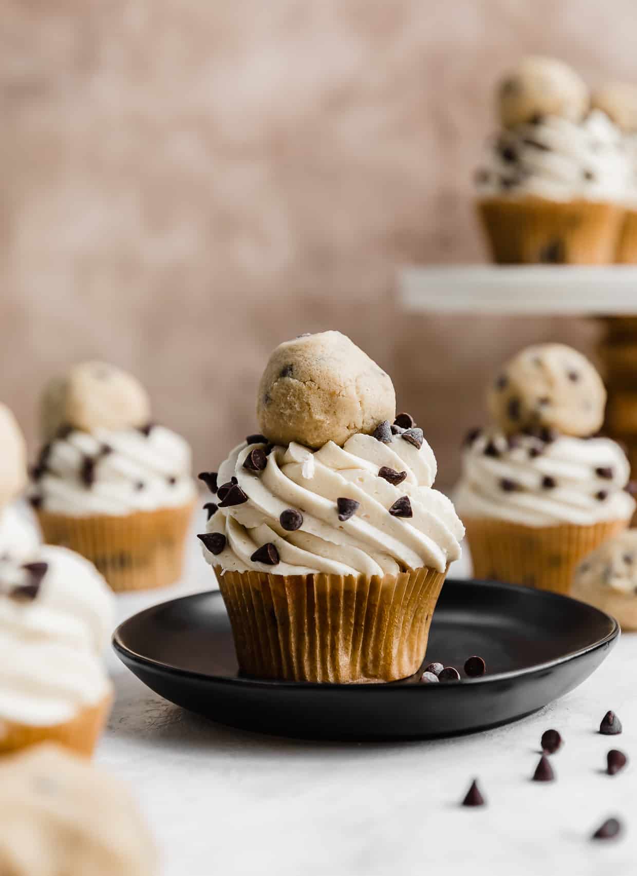 A chocolate chip cookie dough cupcake topped with a swirl of white frosting, topped with mini chocolate chips and a ball of edible cookie dough on top.