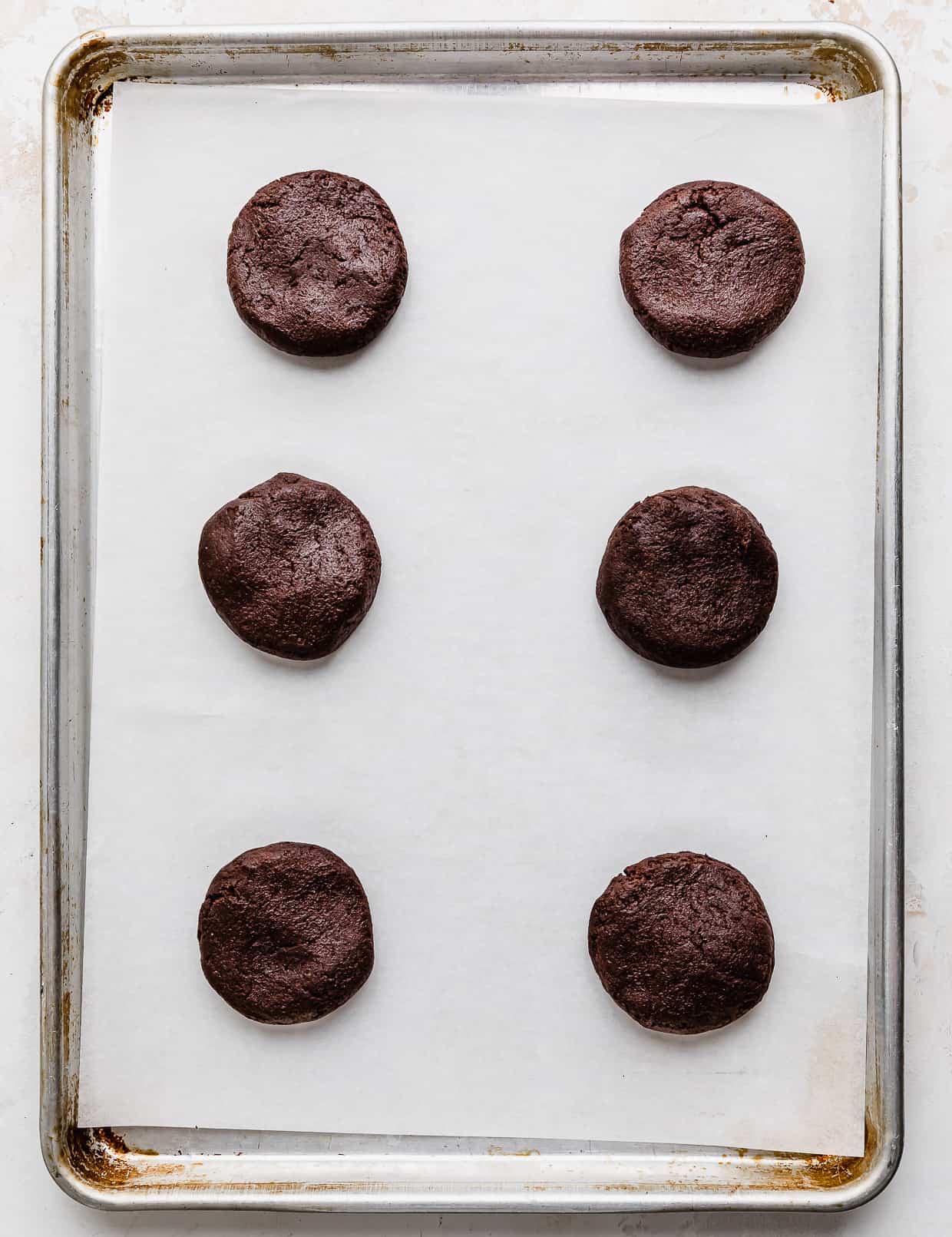 Six chocolate cookie dough balls on a white parchment paper lined baking sheet.