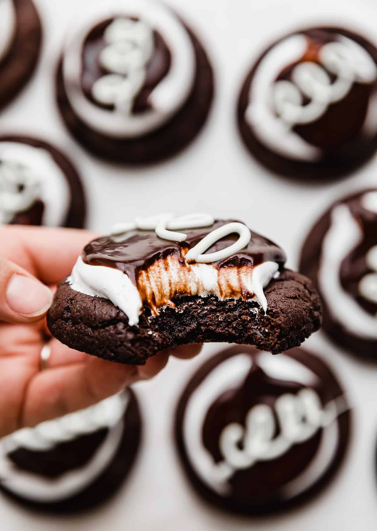 A hand holding a Hostess Cupcake Cookie (chocolate cookie topped with marshmallow frosting and chocolate ganache) with a bite taken out of it.