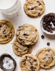 Six chocolate Chip Cookies on a white background with a small bowl filled with sea salt and chocolate chips.