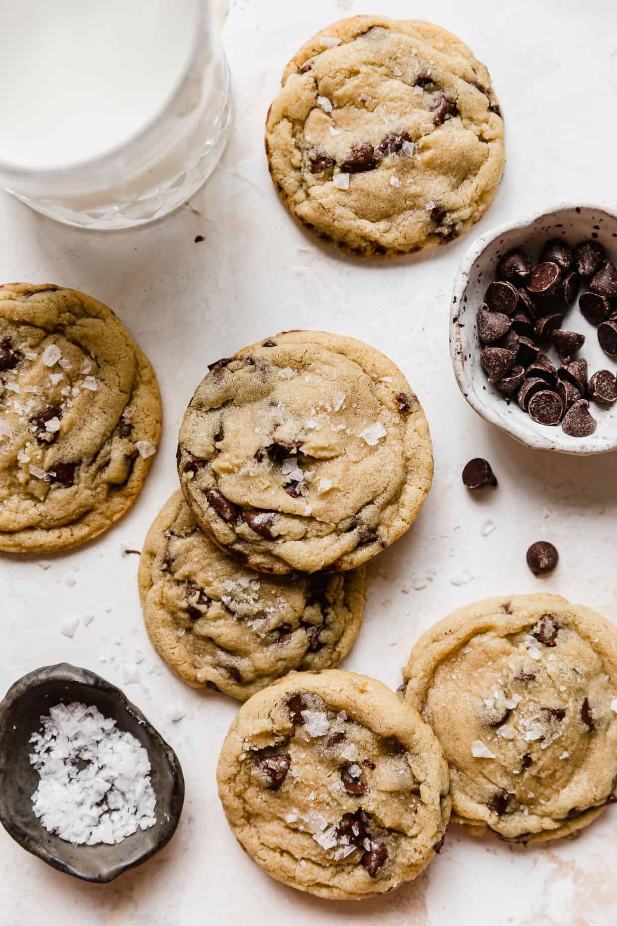 Six chocolate Chip Cookies on a white background with a small bowl filled with sea salt and chocolate chips. 