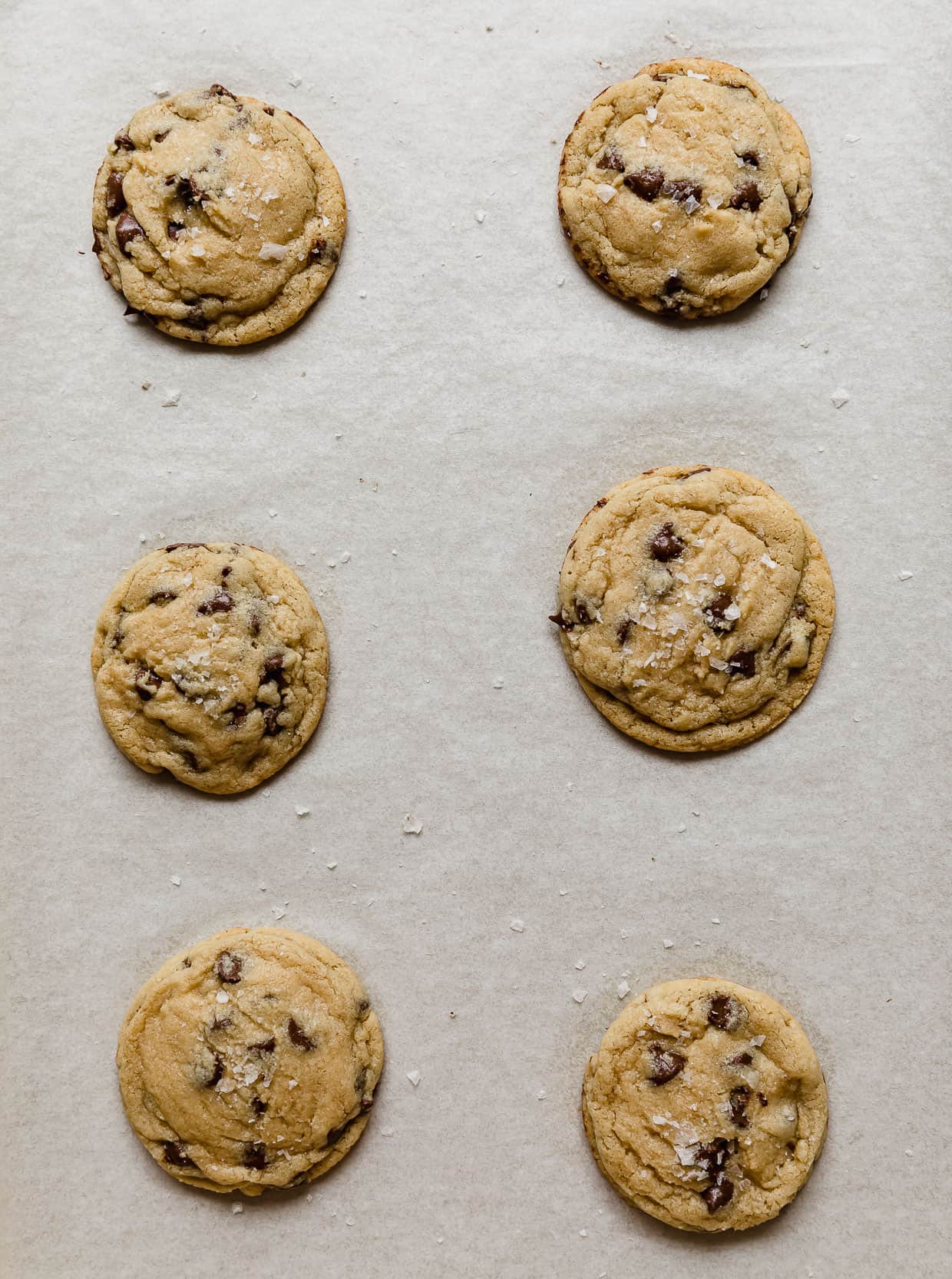 Six baked chocolate chip cookies on a white parchment lined baking sheet.
