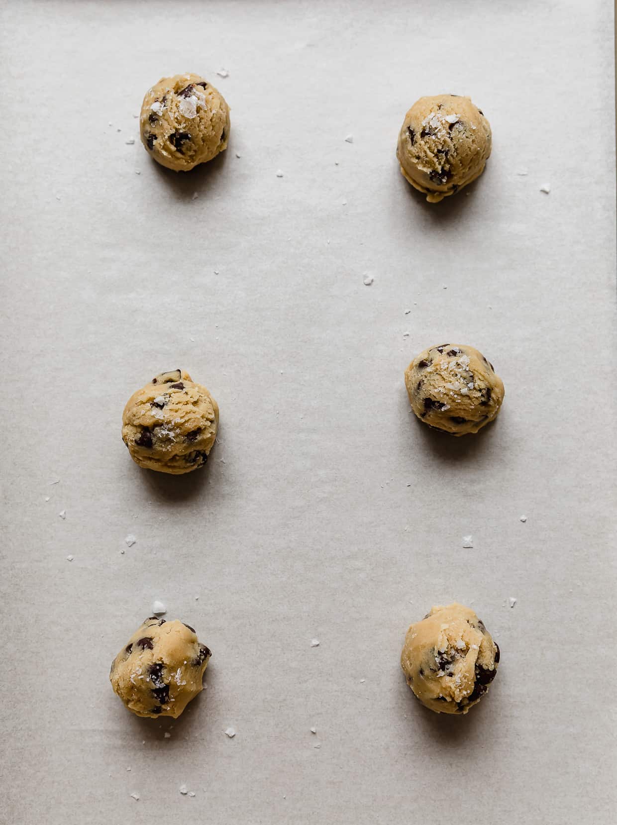 Six chocolate chip cookie dough balls on a white parchment lined baking sheet.