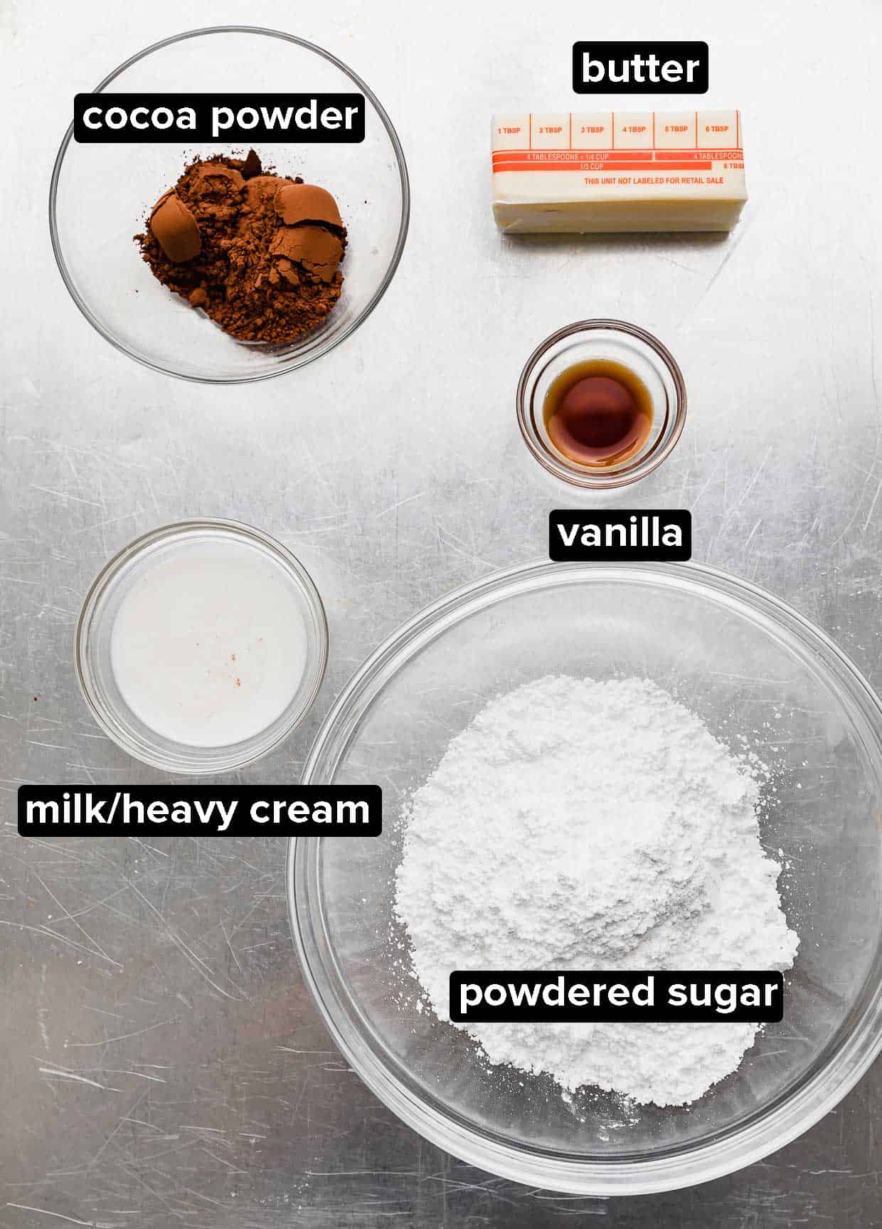Cocoa powder, powdered sugar, milk, vanilla, and butter on a silver baking sheet.