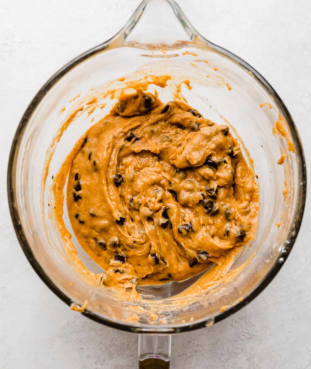 A glass mixing bowl filled with Crumbl Pumpkin Chocolate Chip Cookie batter on a white background. 