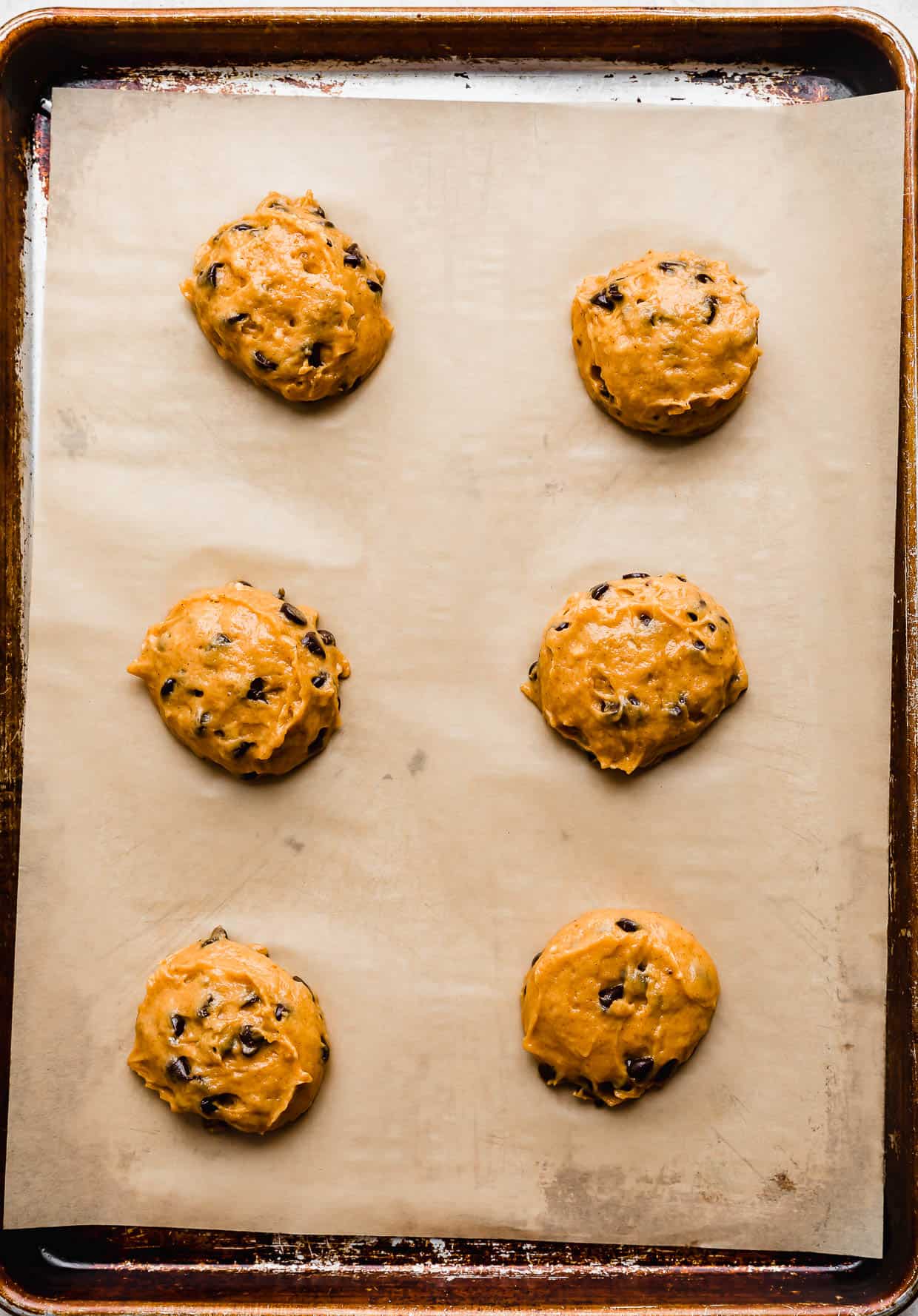 Six balls of Crumbl Pumpkin Chocolate Chip Cookie dough batter on a tan parchment lined baking sheet.