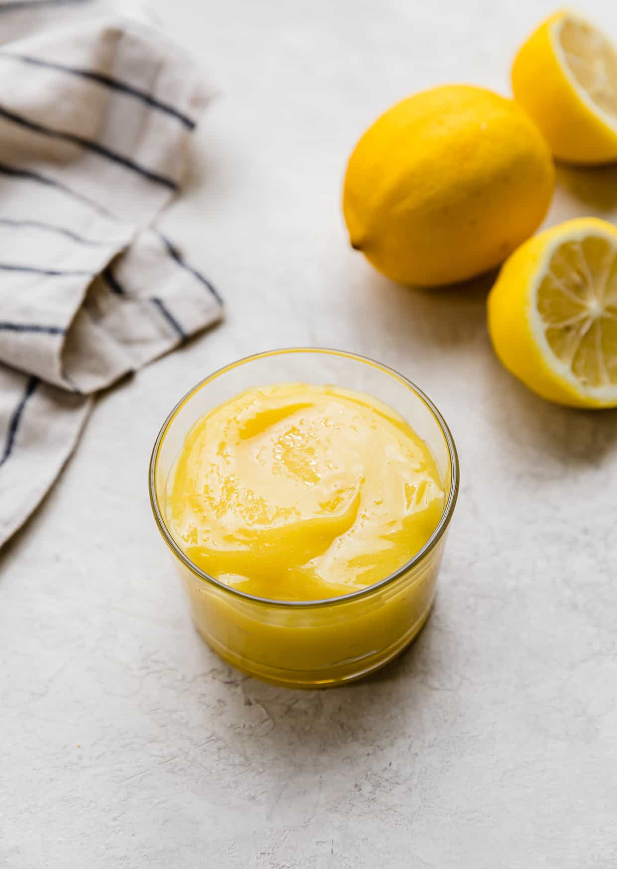 A homemade Lemon Curd Recipe in a glass cup on a light gray background with a full lemon and two lemon halves in the background.