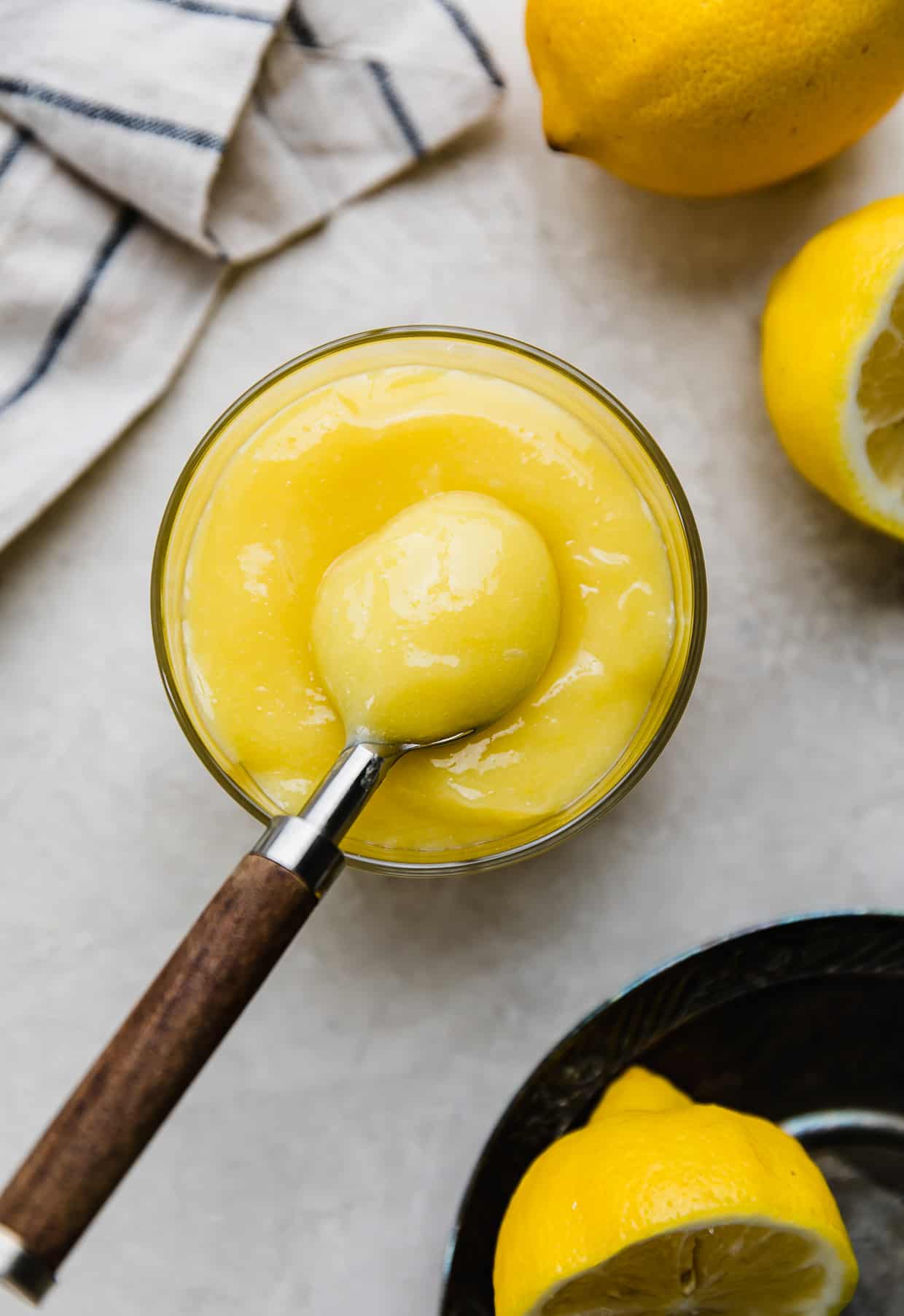 Overhead photo of yellow lemon curd in a glass cup on a light gray background, with a brown spoon scooping up a portion of the lemon curd.