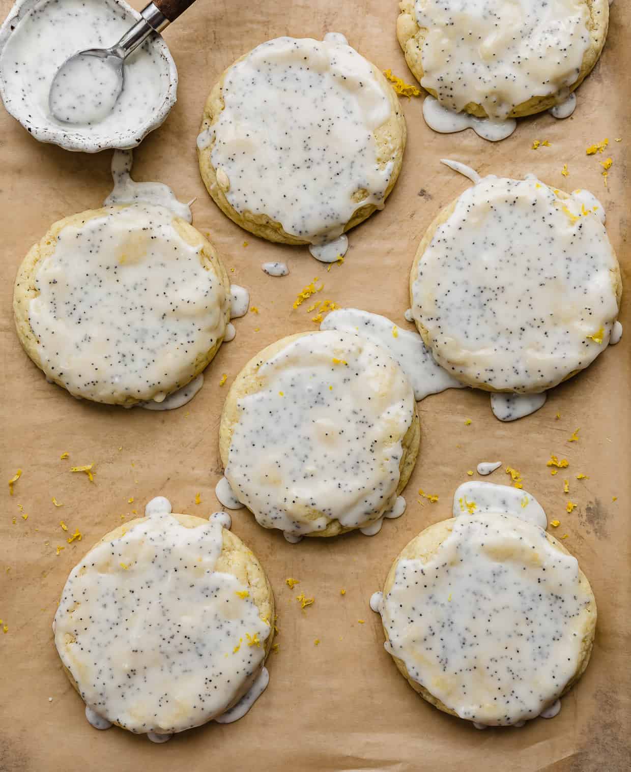 A lemon almond poppy seed glaze overtop of lemon poppy seed cookies that are on a tan colored parchment paper.