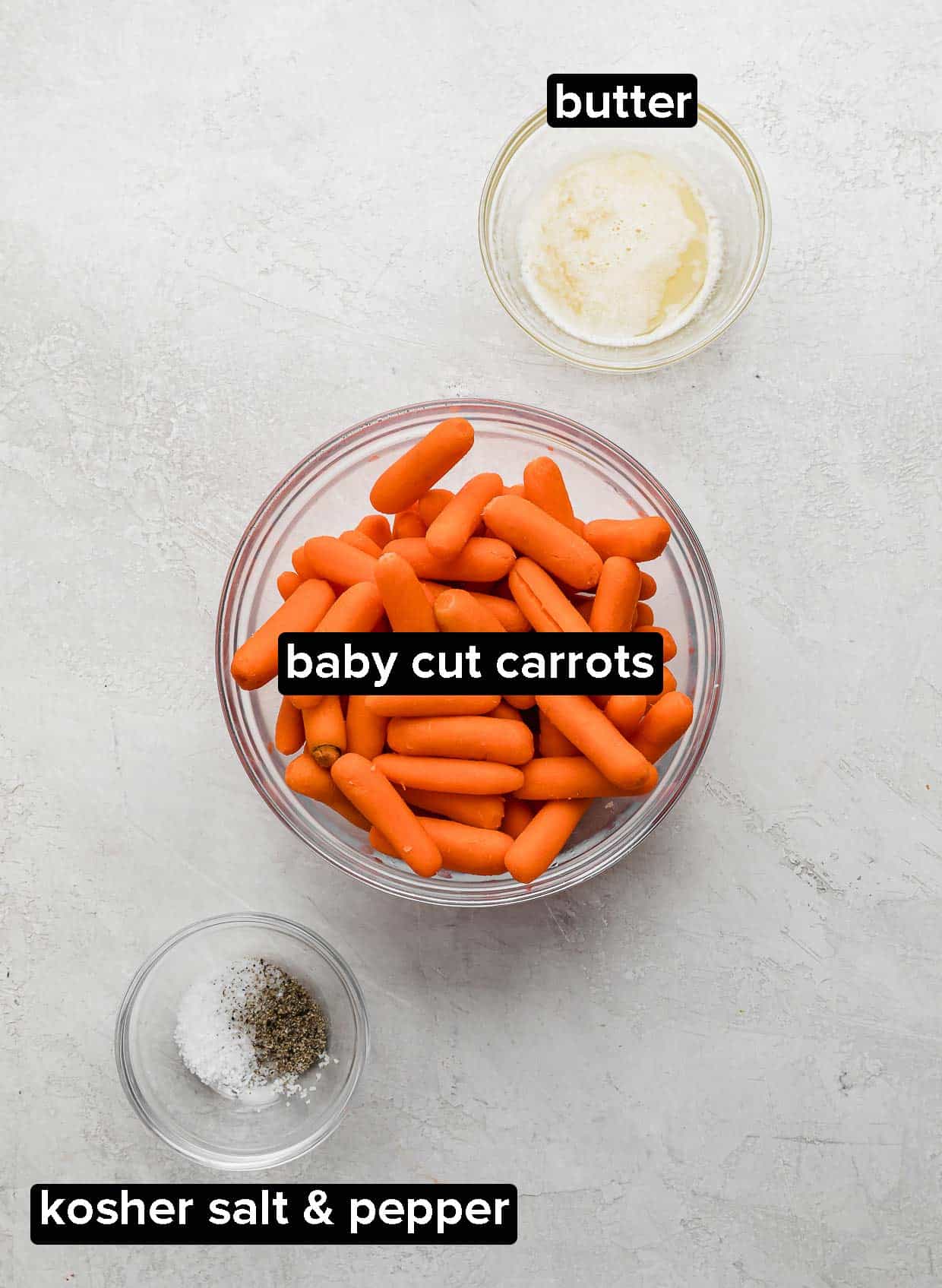 A white background with baby carrots in a glass bowl, butter, and salt and pepper.