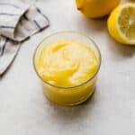 Homemade Lemon Curd in a glass cup on a light gray background with a lemon half in the background.