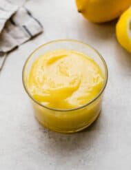 Homemade Lemon Curd in a glass cup on a light gray background with a lemon half in the background.