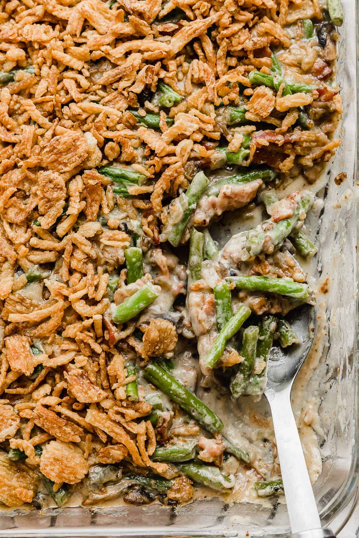 A serving spoon scooping some Bacon Green Bean Casserole topped with French fried onions in a baking dish.