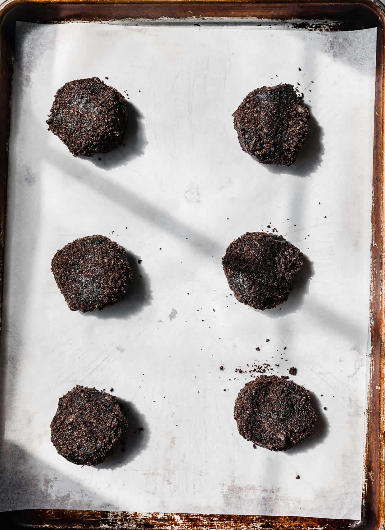 Six black Dirt Cake Cookie dough balls on a white parchment lined baking sheet.