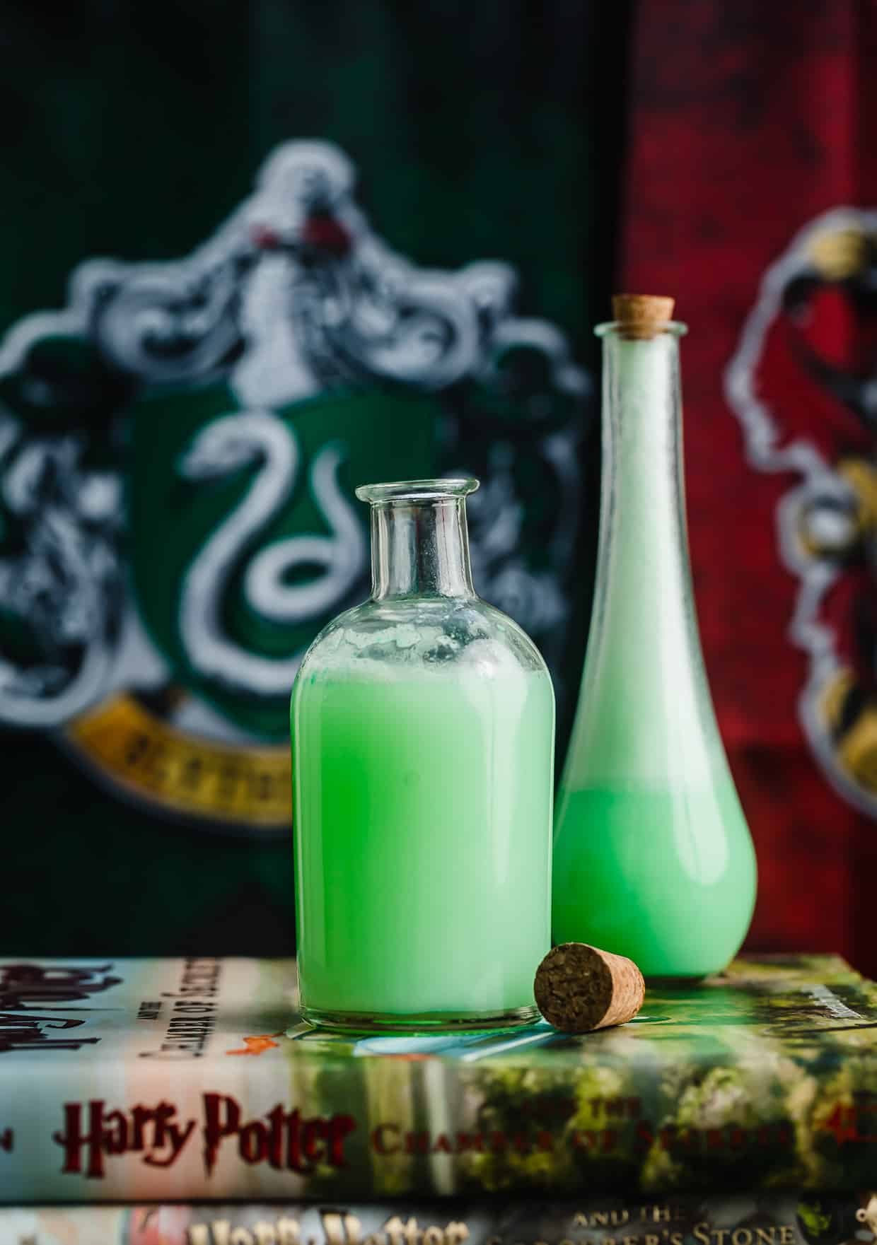 Two potion jars filled with a green Polyjuice Potion drink that's on top of a Harry Potter Chamber of Secrets book.