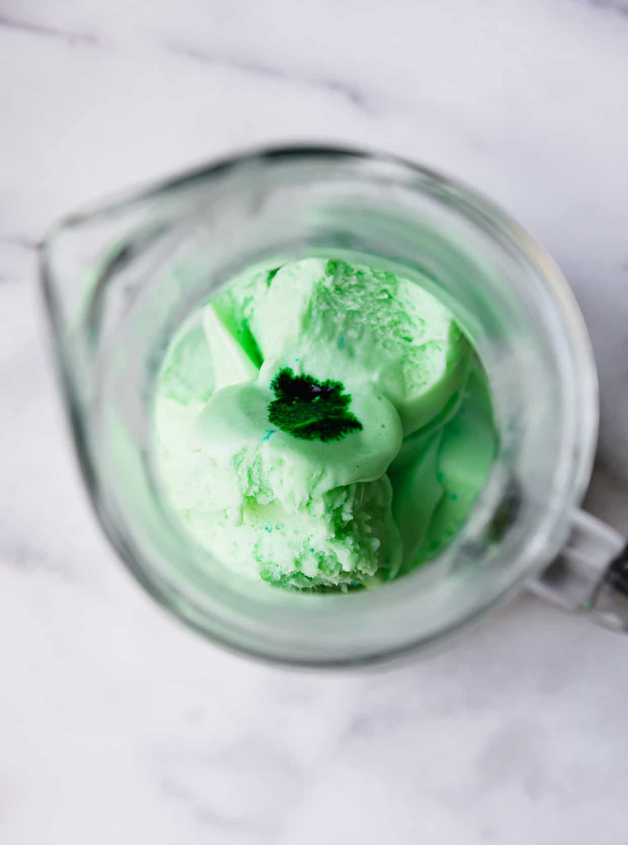 Overhead photo of lime sherbet and a drop of green food color in a glass pitcher.