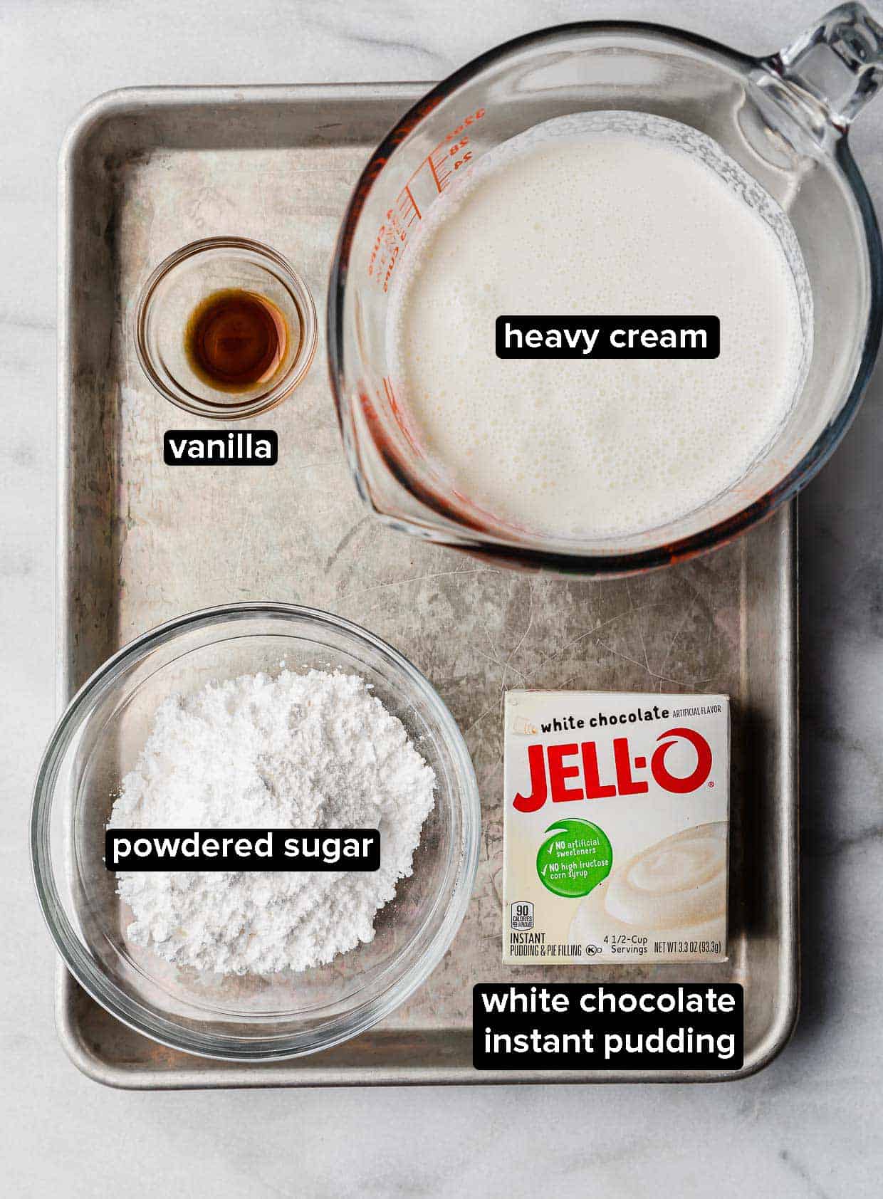 Ingredients used to make Whipped Cream Frosting with pudding on a white background: heavy cream, vanilla, powdered sugar, and white chocolate instant pudding.