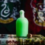 Green Harry Potter Polyjuice Potion in a glass potion bottle in front of a Slytherin and Gryffindor house flag.