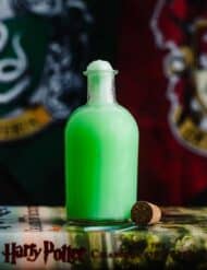 Green Harry Potter Polyjuice Potion in a glass potion bottle in front of a Slytherin and Gryffindor house flag.