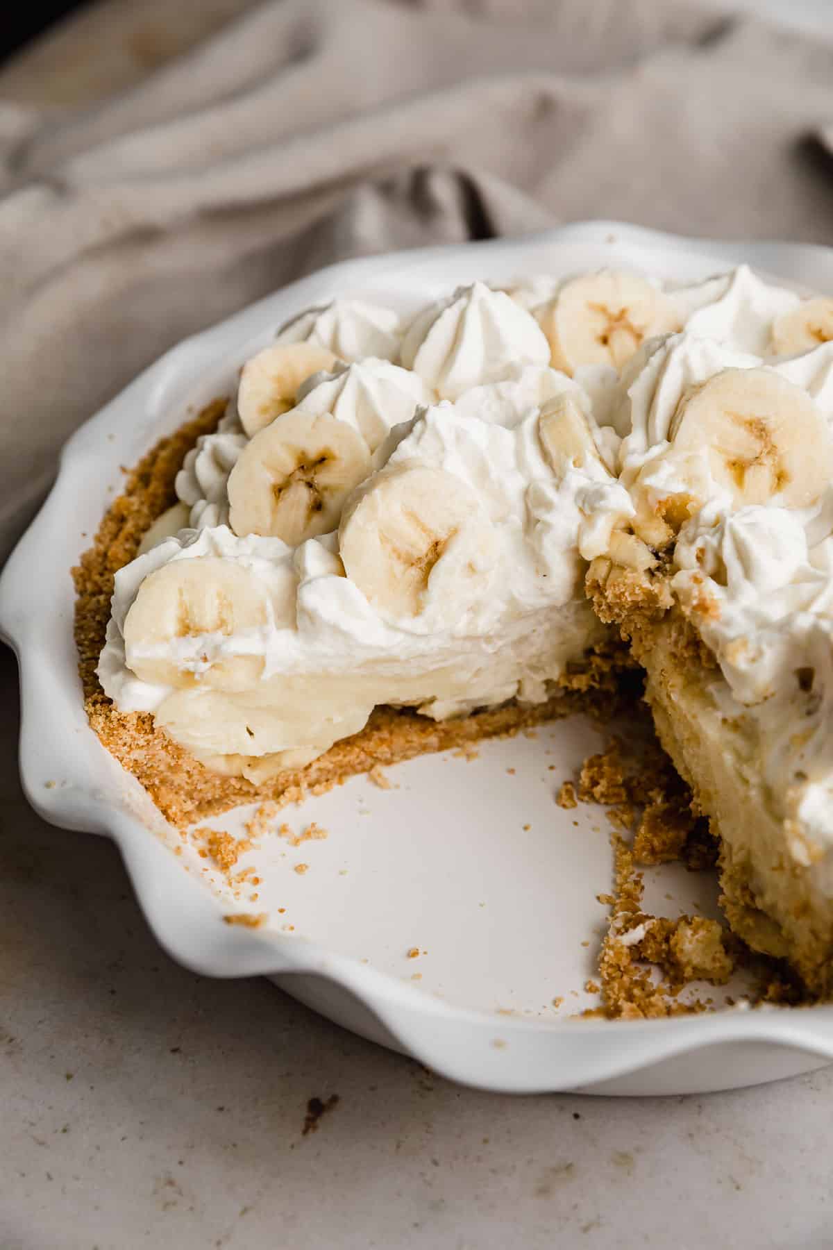 A banana cream pie in a white pie plate with a slice removed from the pie showing the sliced bananas underneath the custard.