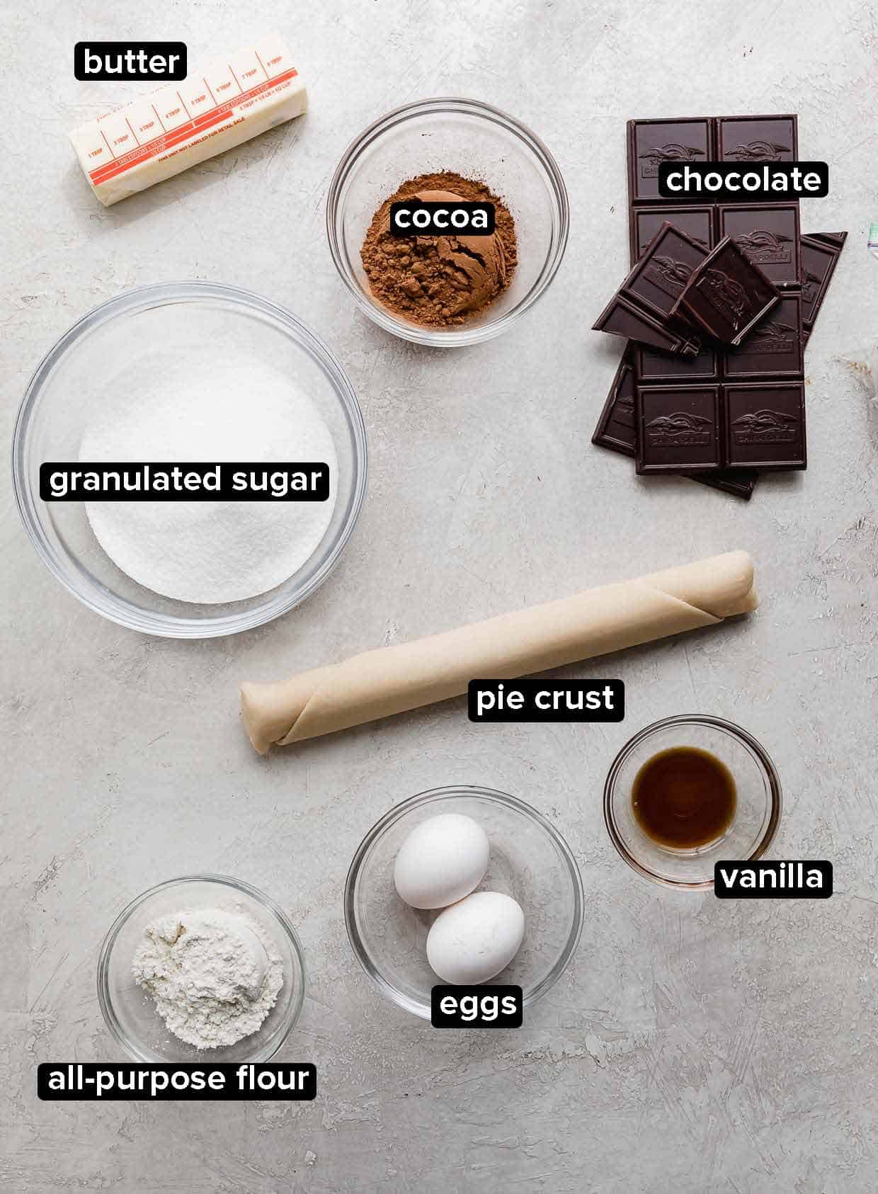 Brownie Pie ingredients on a light gray background, including chocolate, cocoa, pie crust, eggs, vanilla, butter, and sugar.