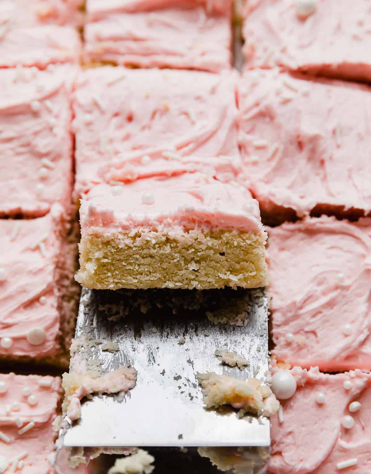 A metal spatula lifting up a square of Sugar Cookie Bar that's frosted with pink buttercream frosting.