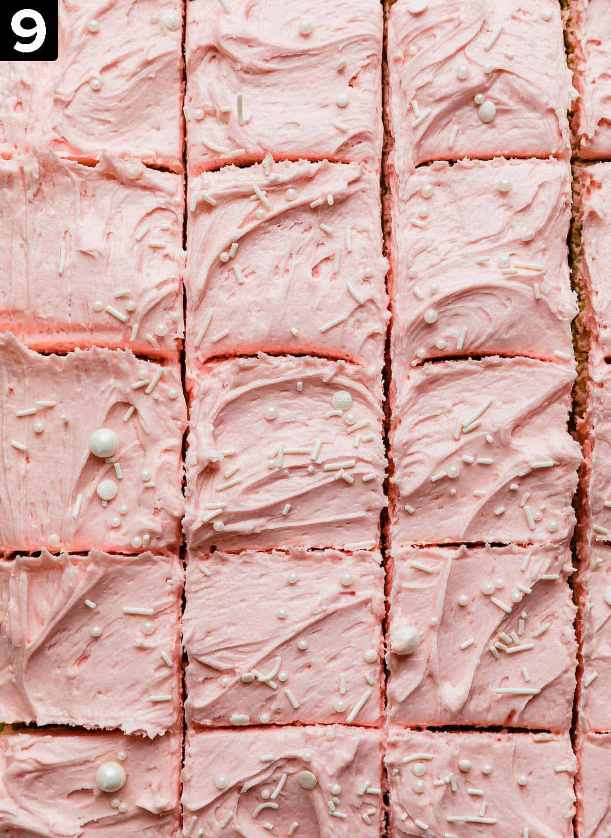 Pink frosting covered Sugar Cookie Bars topped with a sprinkling of white sprinkles, and the bars are cut into small squares.