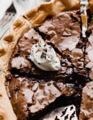 A brownie pie sliced into several servings, with one slice topped with white whipped cream and chocolate shavings.