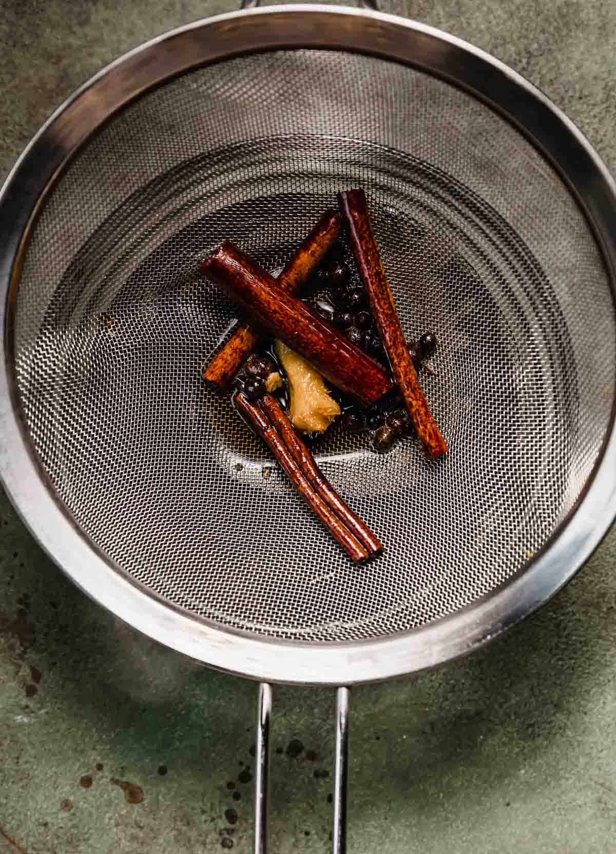 A metal strainer with four cinnamon sticks, handful of allspice berries, and small fresh ginger chunk in it.