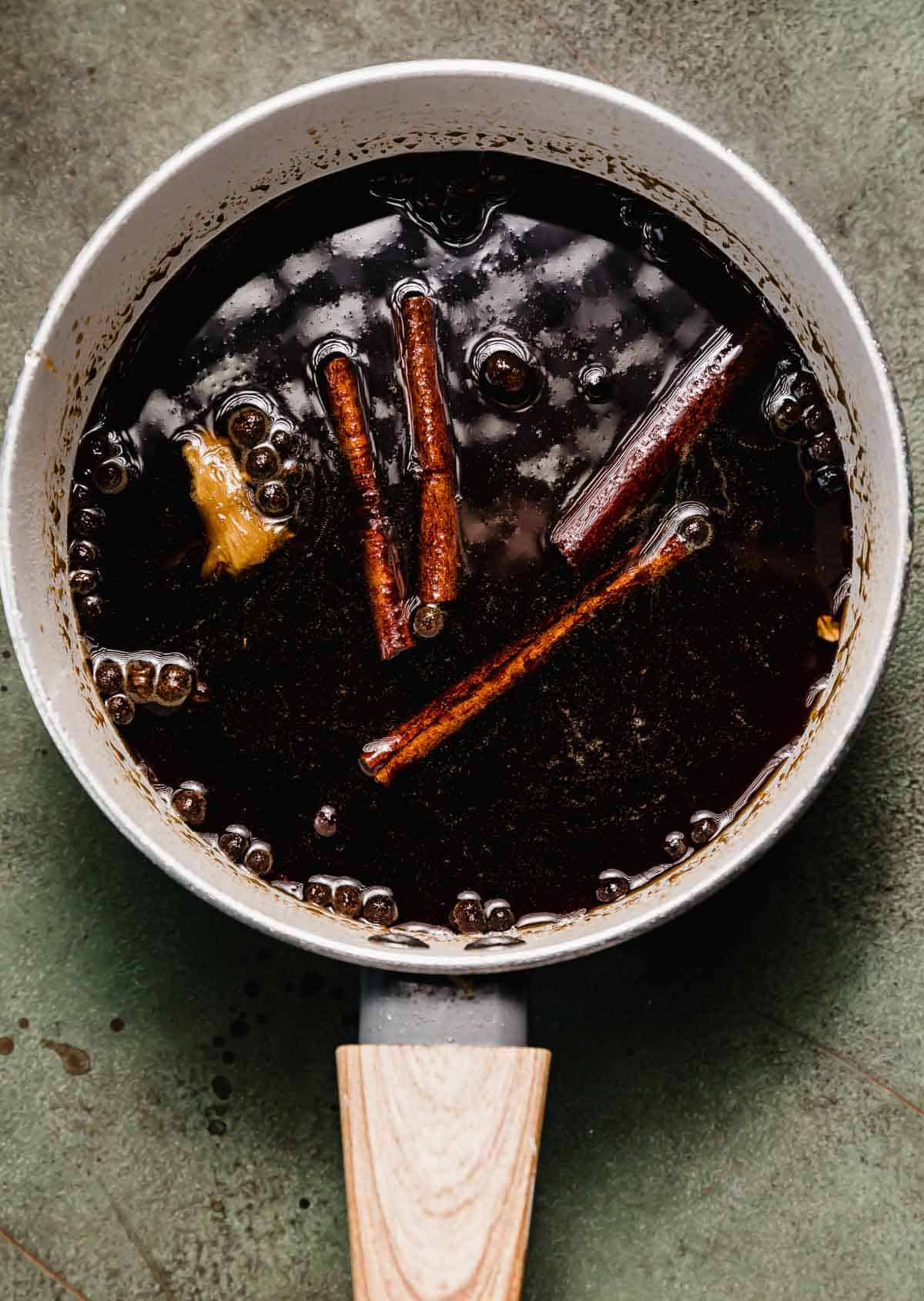 Allspice berries, cinnamon sticks, and fresh ginger in a dark Gingerbread Syrup that's in a saucepan on a green background.