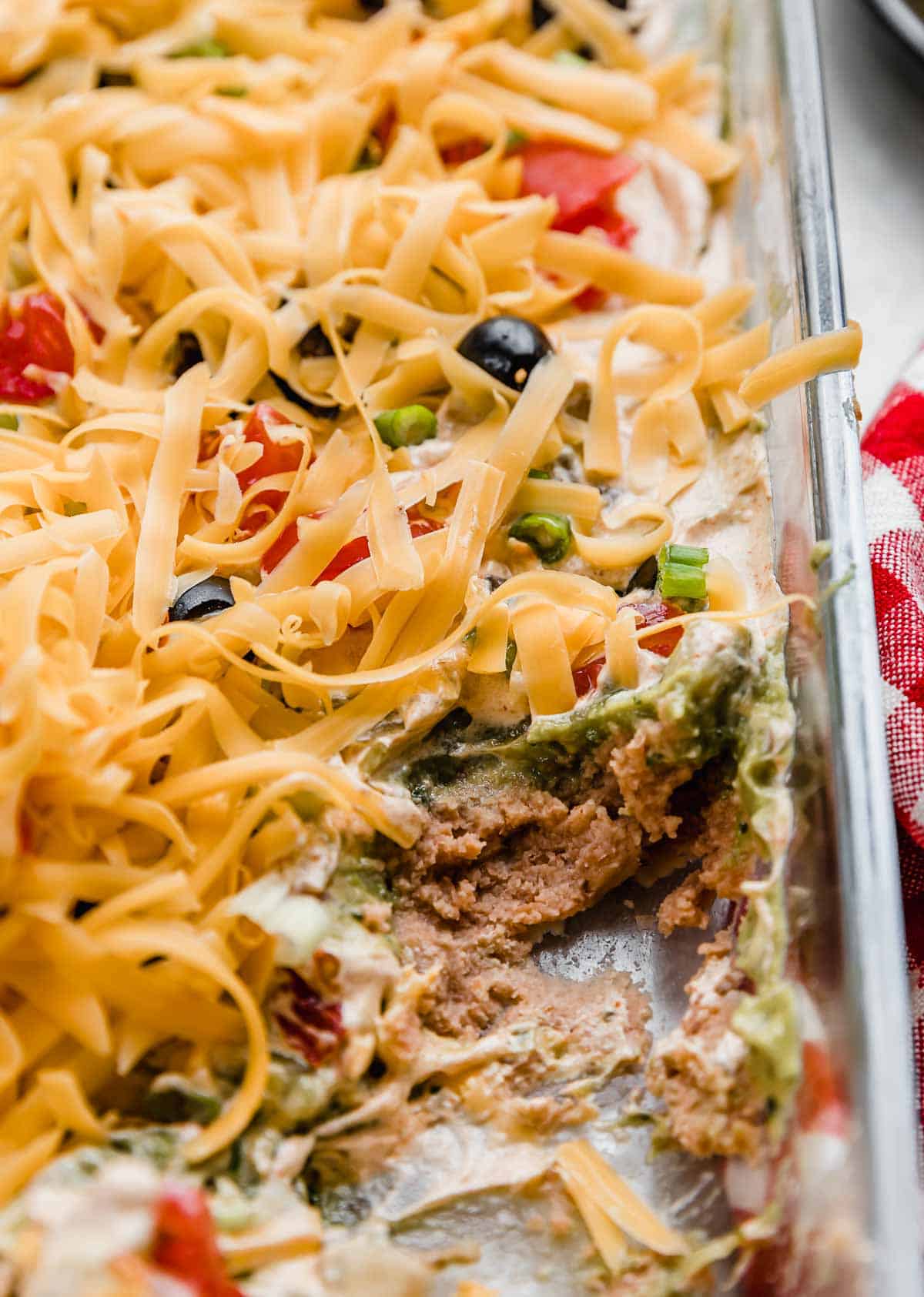 Layered Taco Dip in a glass baking dish, refried beans, guacamole, tomatoes, and cheddar cheese topped dip.