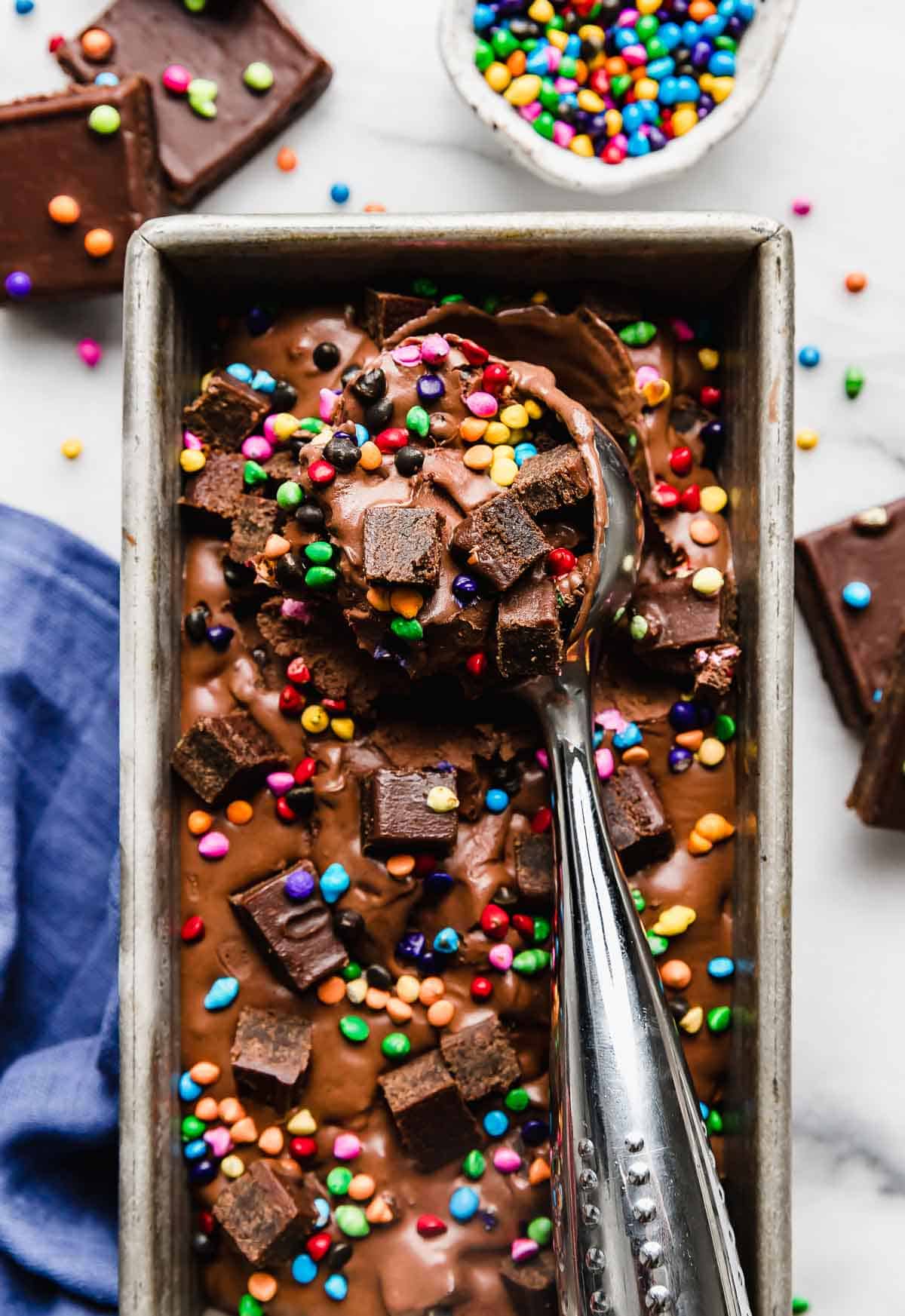 An ice cream scoop scooping out Cosmic Brownie Ice Cream from a rectangular bread pan on a white marble background.
