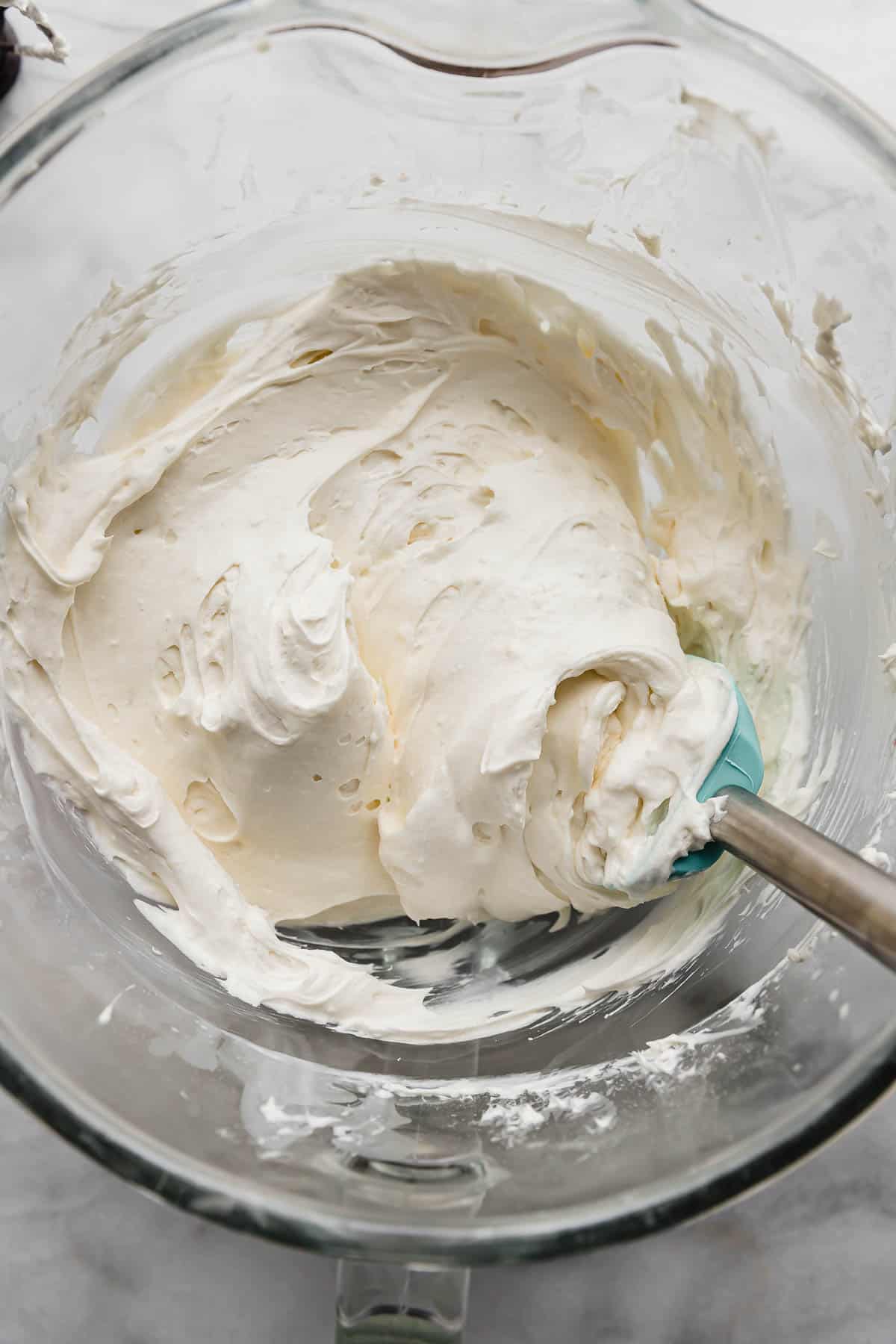 A turquoise blue rubber spatula in a bowl filled with white Cream Cheese Whipped Cream Frosting.