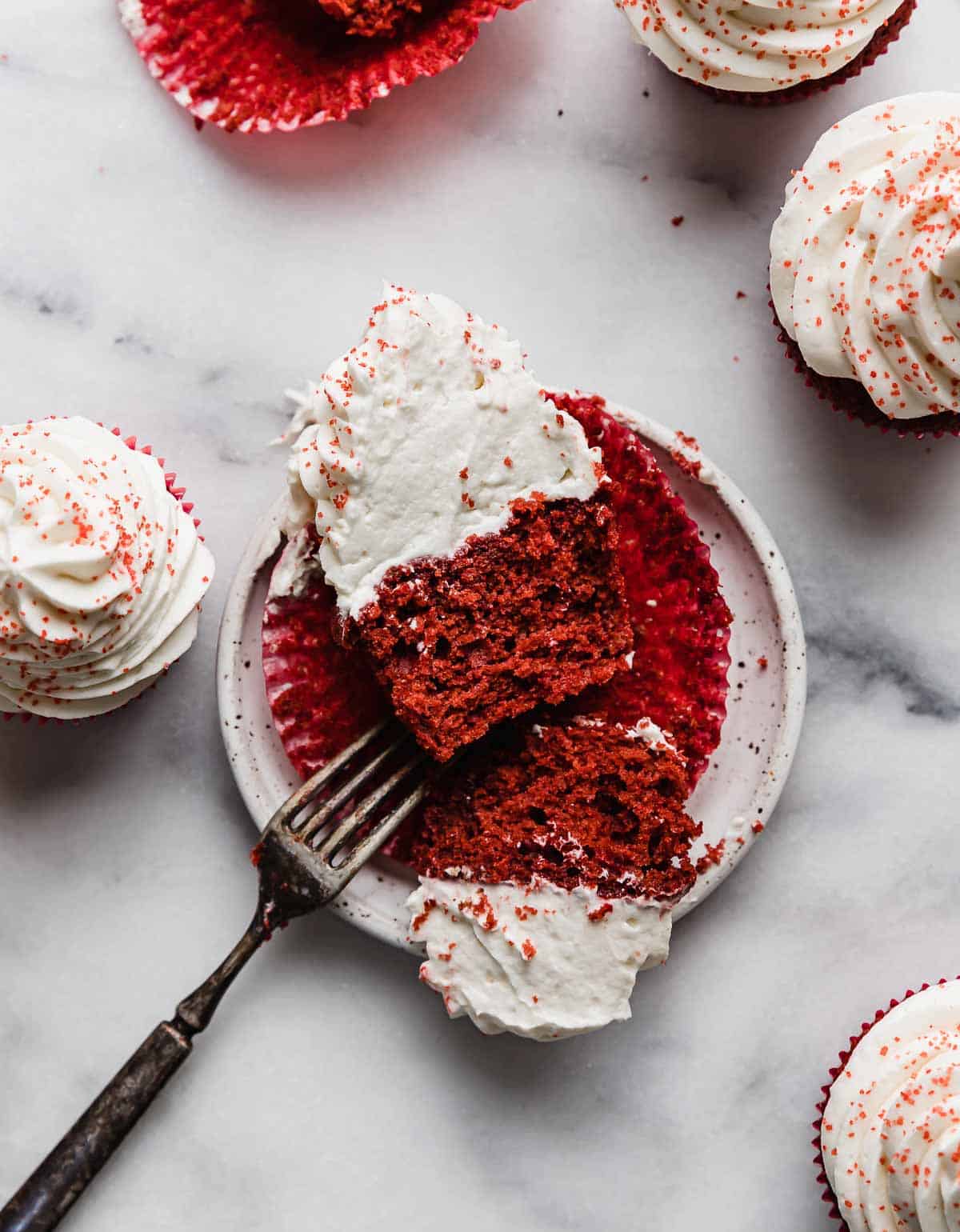 A Red Velvet Cupcake cut in half, on a white plate that's on a white marble table.