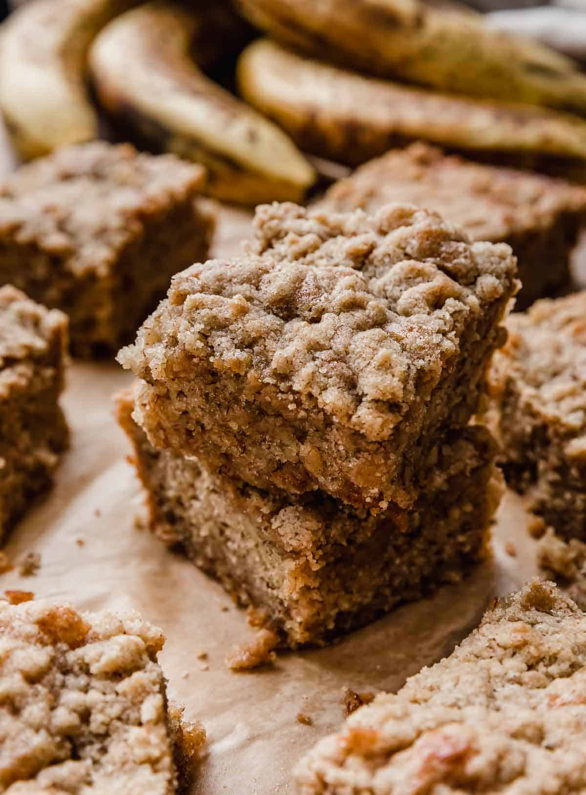 Two squares of moist banana coffee cake stacked on top of each other with ripe bananas in the background. 