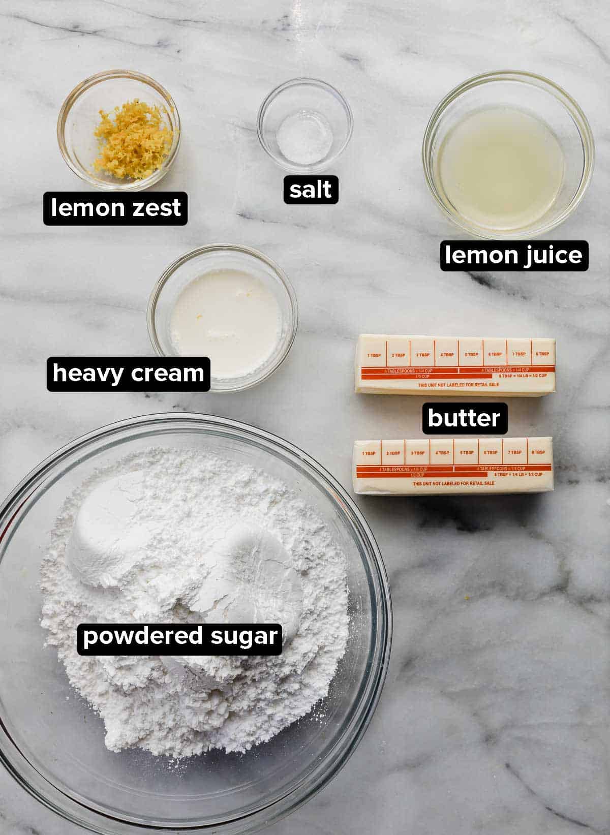 Lemon Buttercream frosting ingredients portioned into glass bowls on a white marble table.