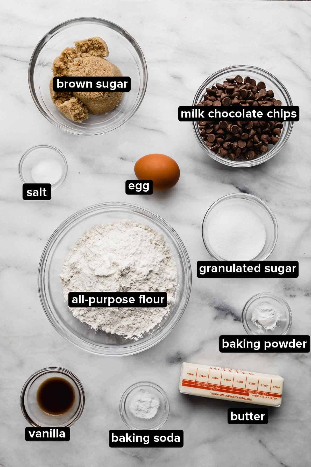 Milk Chocolate Chip Cookie ingredients portioned into glass bowls on a white marbled background.