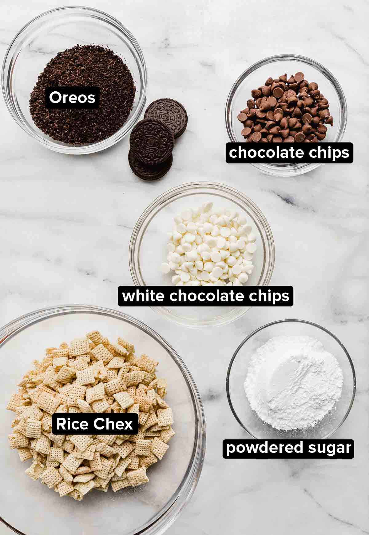 Oreo Muddy Buddies ingredients on a white marble background: Oreo crumbs, Chex cereal, white and milk chocolate chips, and powdered sugar.