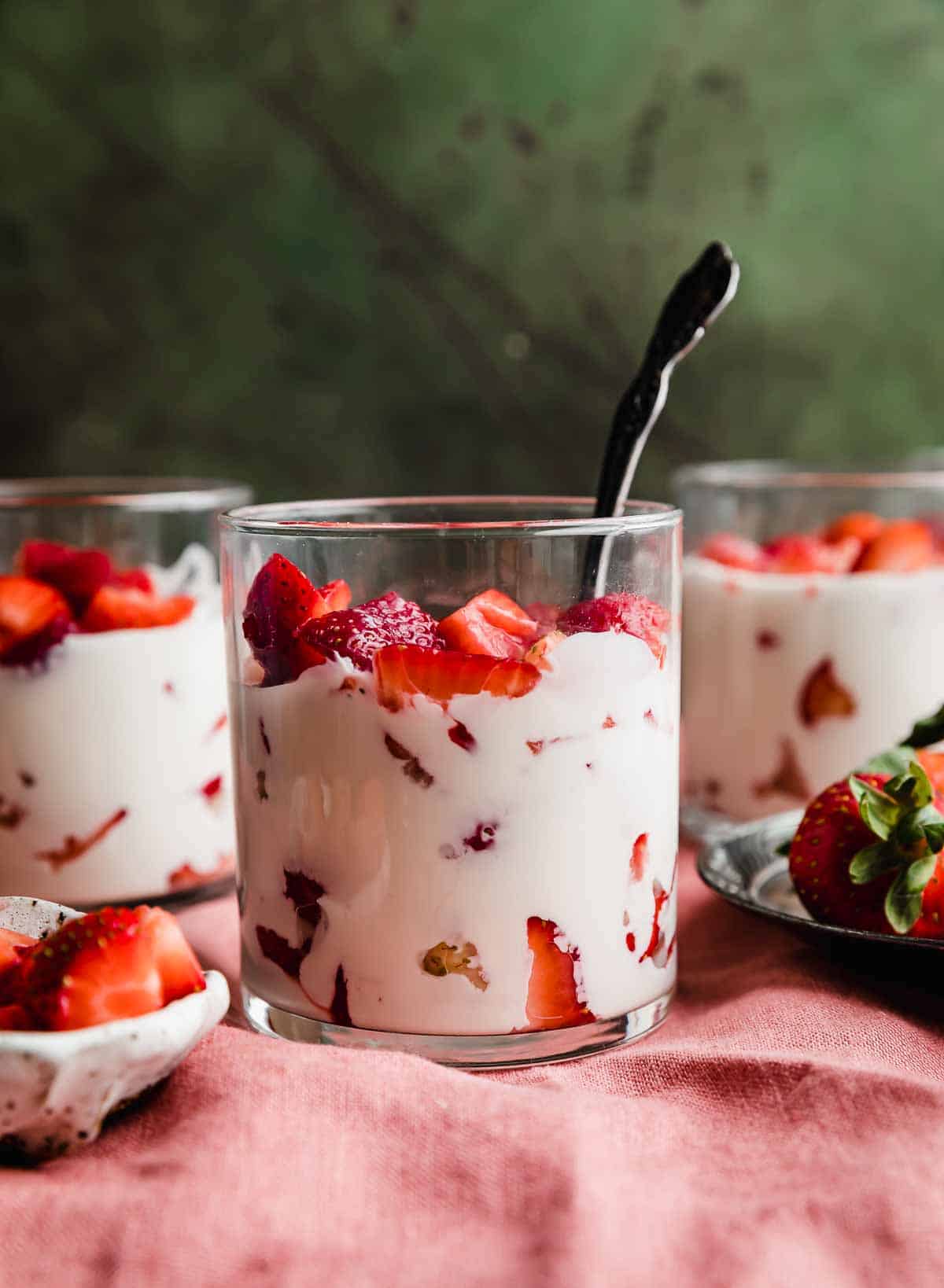 Three clear cups filled with Strawberries and Cream (Fresas con Crema) on a red linen cloth against a textured brown and green background.