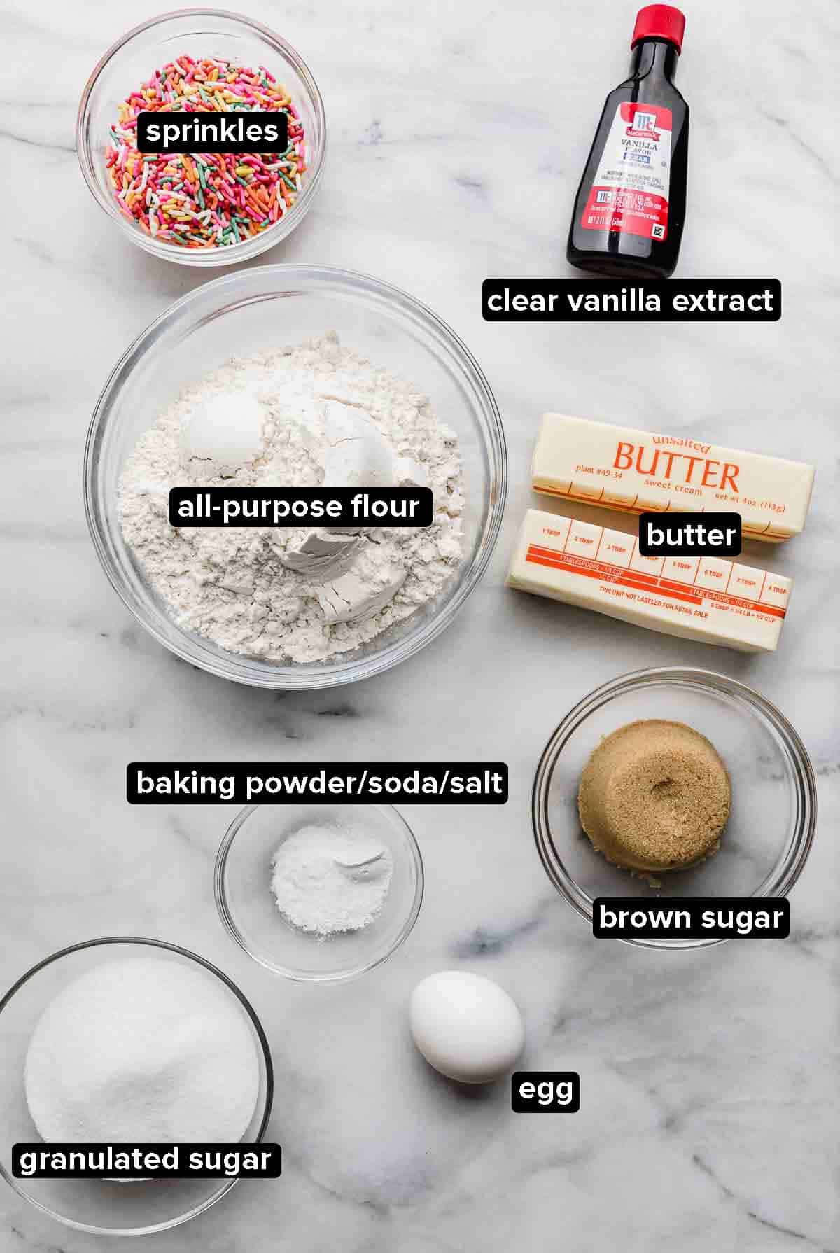 Funfetti Cookie ingredients portioned into glass bowls on a white background: clear vanilla extract, sprinkles, flour, sugar and brown sugar, butter, egg, and leavening agent.