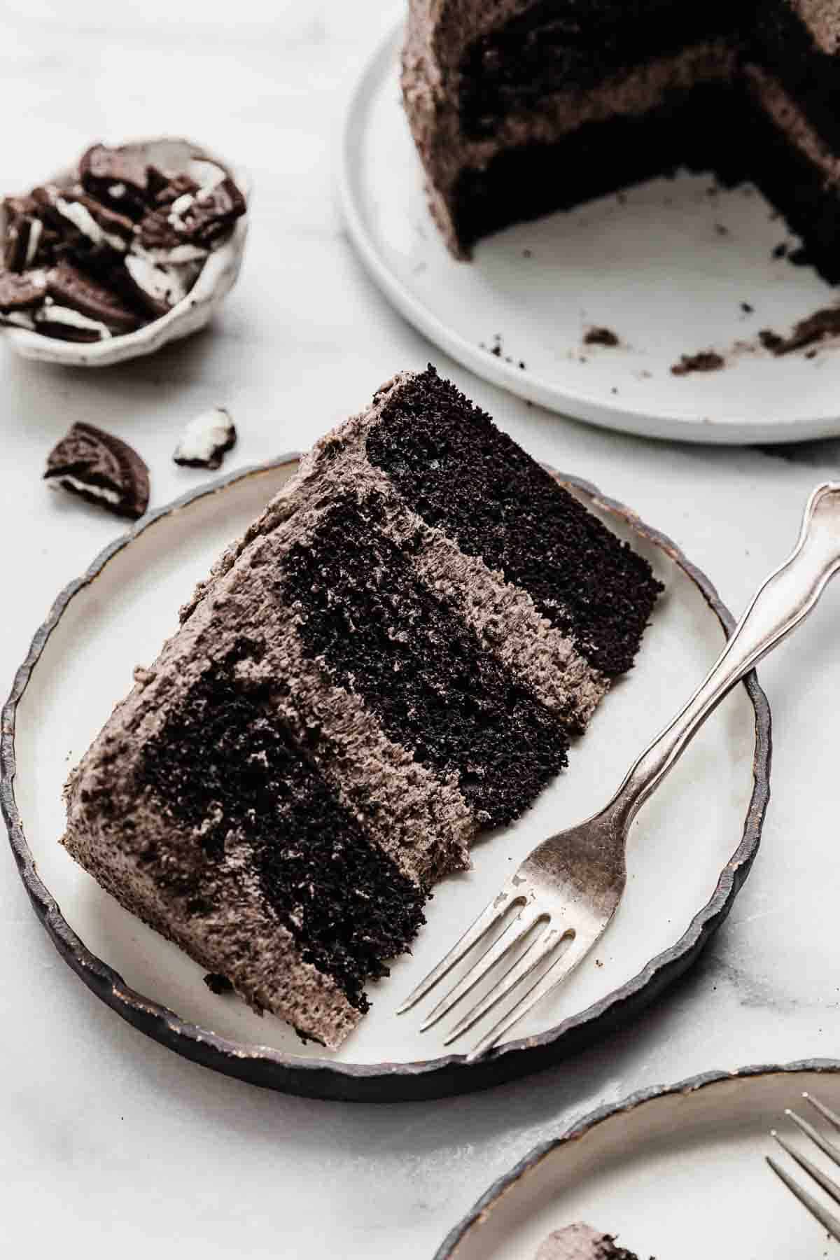 A slice of three layer Oreo Cake frosted with an Oreo frosting, on a black rimmed white plate.