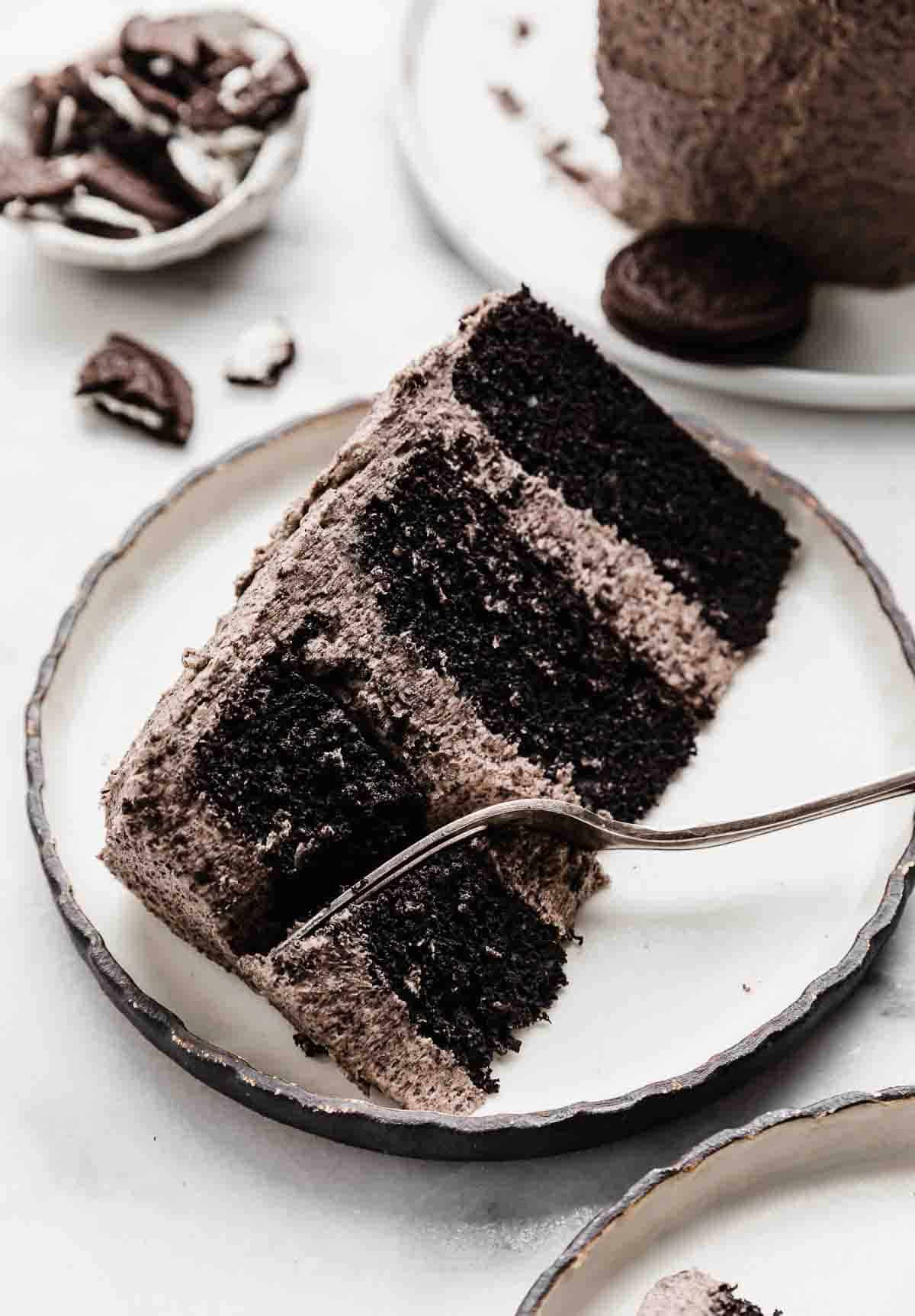 A slice of three tiered oreo cake on a black rimmed white plate with a fork cutting into the top portion of the cake.