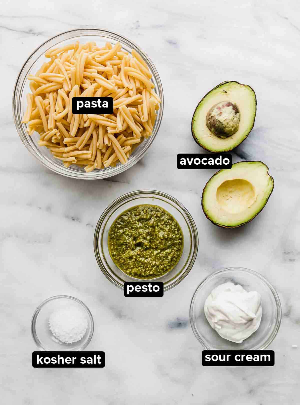 Avocado Pesto Pasta ingredients on a white and gray marble background: noodles, sour cream, salt, basil pesto, and an avocado.