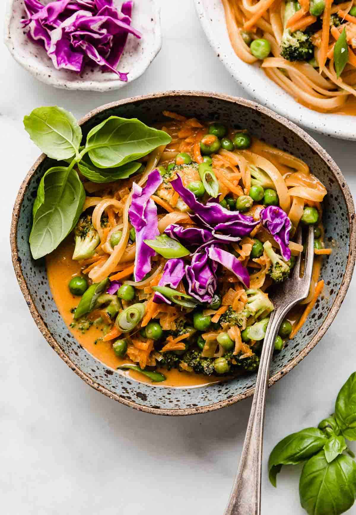 A blue bowl filled with Coconut Curry Noodles and vegetables topped with basil and purple cabbage.