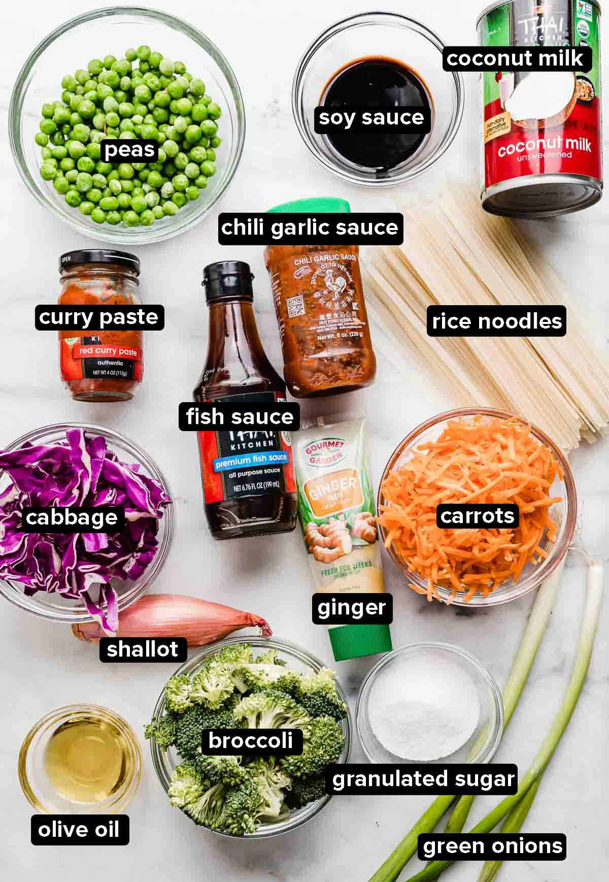 Coconut Curry Noodle Bowls ingredients on a white background: broccoli, rice noodles, curry paste, peas, carrots, purple cabbage, soy sauce, coconut milk.