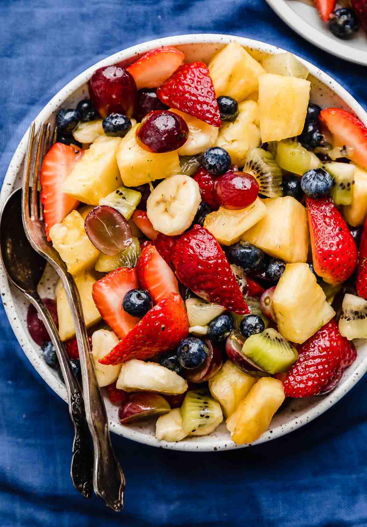 Fruit Cocktail consisting of chopped strawberries, kiwi, pineapple, banana, blueberries on a white plate on a dark blue background.